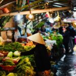 Doc. Seorang pedagang dan pembeli bertransaksi di pasar tradisional yang dipenuhi berbagai jenis sayuran segar. Suasana pasar tampak ramai dengan pencahayaan lampu sederhana, menciptakan nuansa khas pasar rakyat. Seorang perempuan mengenakan caping tampak memilih sayuran, sementara pedagang lain sibuk menata dagangannya. Gambar ini hanya merupakan ilustrasi dan tidak secara spesifik menggambarkan kondisi pasar tertentu. (Gambar: NR).