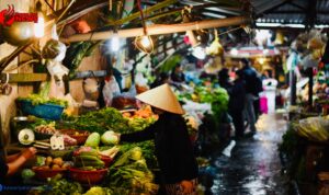 Doc. Seorang pedagang dan pembeli bertransaksi di pasar tradisional yang dipenuhi berbagai jenis sayuran segar. Suasana pasar tampak ramai dengan pencahayaan lampu sederhana, menciptakan nuansa khas pasar rakyat. Seorang perempuan mengenakan caping tampak memilih sayuran, sementara pedagang lain sibuk menata dagangannya. Gambar ini hanya merupakan ilustrasi dan tidak secara spesifik menggambarkan kondisi pasar tertentu. (Gambar: NR).