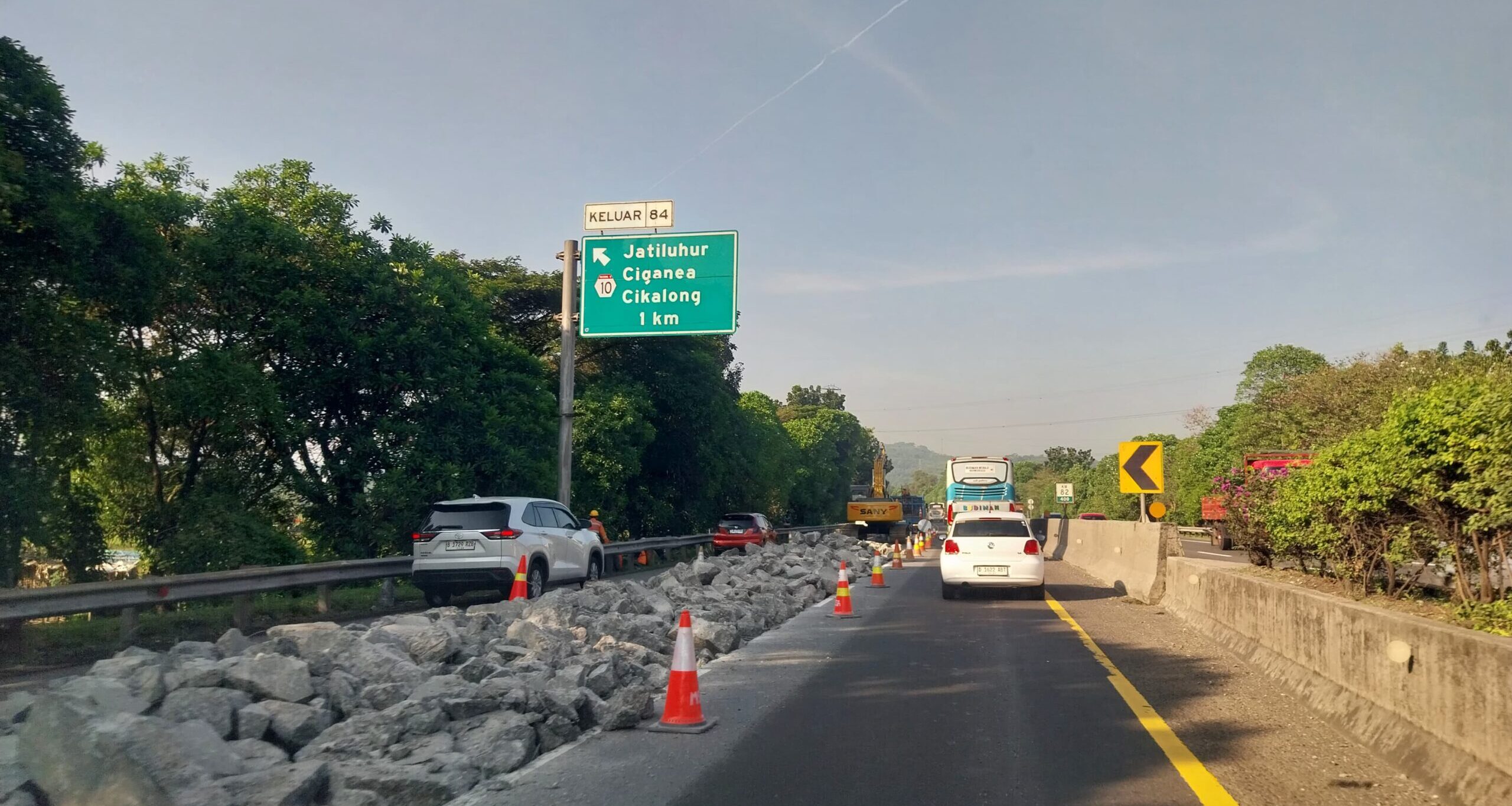 Doc. Pekerjaan konstruksi tengah berlangsung di bahu jalan tol, dengan alat berat dan tumpukan batu besar di sisi kiri. Terlihat papan penunjuk arah keluar KM 84 menuju Jatiluhur, Ciganea, dan Cikalong. Lalu lintas sedikit tersendat karena penyempitan lajur. (Foto Dokumentasi: Nanda).