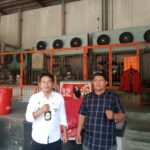 Doc. Sukindar (kiri) berpose bersama seorang rekan di area fasilitas pendingin gudang, dengan latar belakang deretan mesin pendingin industri dan pagar oranye. Keduanya tampak berdiri sambil mengepalkan tangan. (Foto: Ilma).