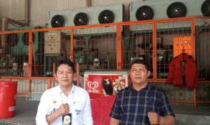 Doc. Sukindar (kiri) berpose bersama seorang rekan di area fasilitas pendingin gudang, dengan latar belakang deretan mesin pendingin industri dan pagar oranye. Keduanya tampak berdiri sambil mengepalkan tangan. (Foto: Ilma).