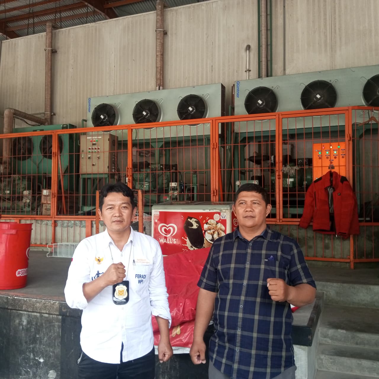 Doc. Sukindar (kiri) berpose bersama seorang rekan di area fasilitas pendingin gudang, dengan latar belakang deretan mesin pendingin industri dan pagar oranye. Keduanya tampak berdiri sambil mengepalkan tangan. (Foto: Ilma).
