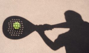 Doc. Ilustrasi bayangan seorang pemain padel yang bersiap memukul bola di lapangan. Pantulan cahaya membentuk siluet raket dan bola di permukaan lapangan, menggambarkan dinamika serta intensitas olahraga padel yang tengah berkembang di kawasan perkotaan. (NR).
